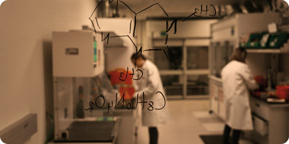 Chemical structure on glass whiteboard with two scientists in the lab blurred in the background