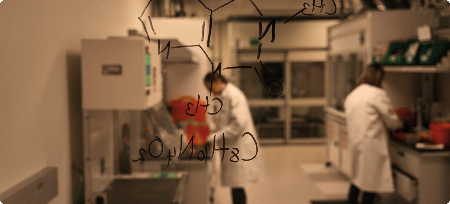 Chemical structure on glass whiteboard with two scientists in the lab blurred in the background